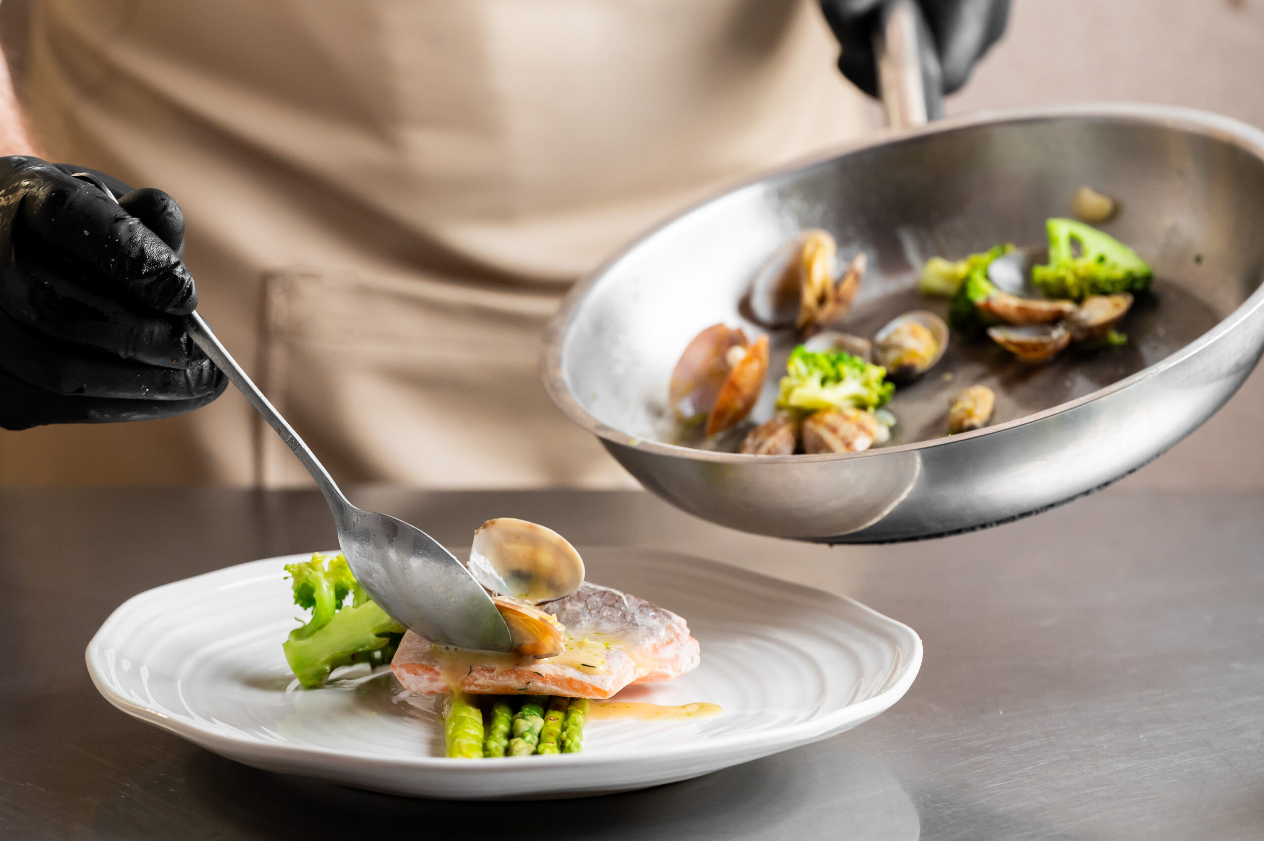 a chef in black gloves uses a spoon to plate cooked clams over a piece of salmon with asparagus and broccoli. a stainless steel pan with more seafood and vegetables is held aloft.