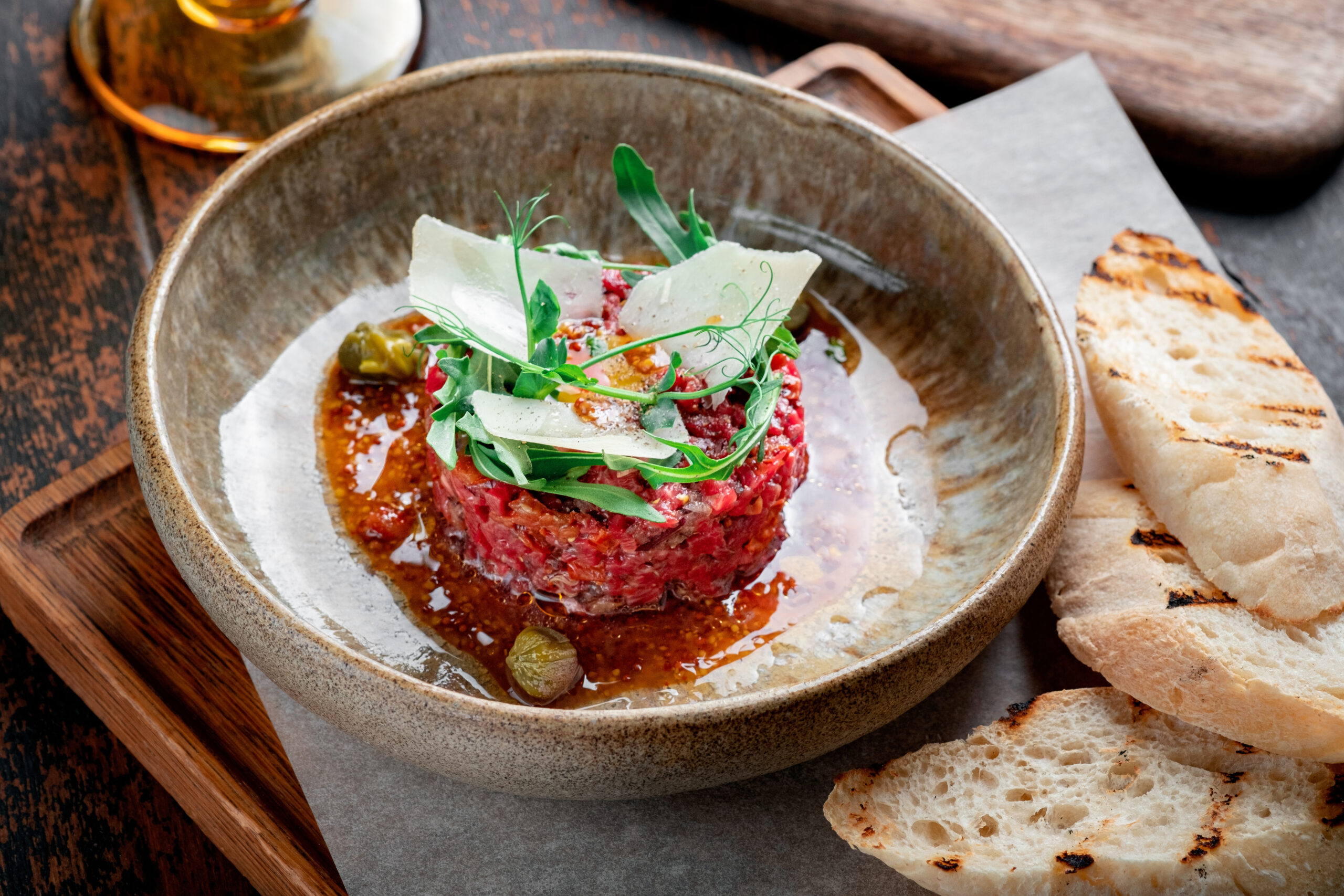 beef tartare with dried tomatoes decorated with parmesan and arugula is served in a plate on a wooden table