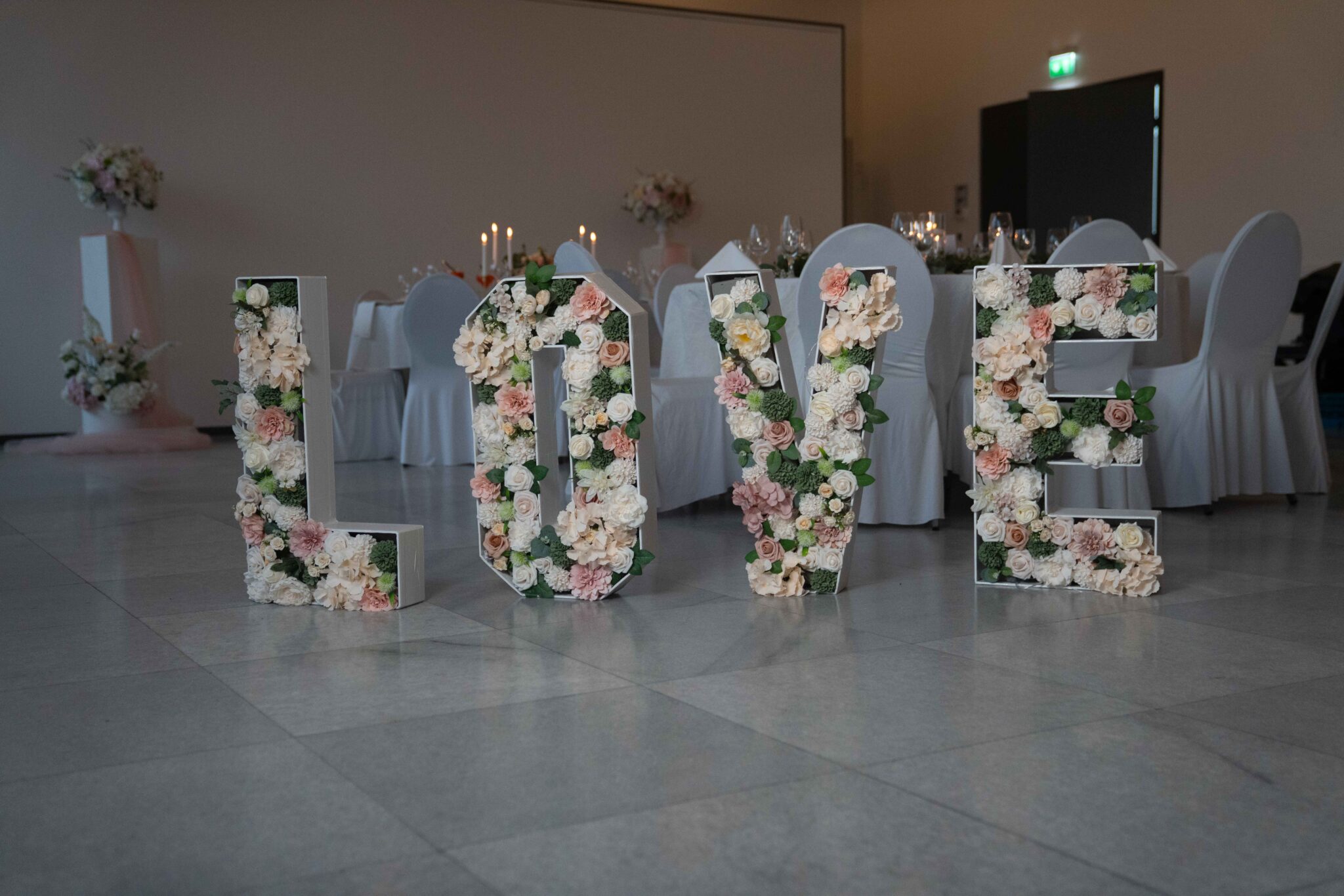 Große, mit Blumen verzierte Blockbuchstaben mit der Aufschrift LOVE" sind auf dem Boden eines dekorierten Veranstaltungsraums mit Tischen und Stühlen im Hintergrund angebracht.