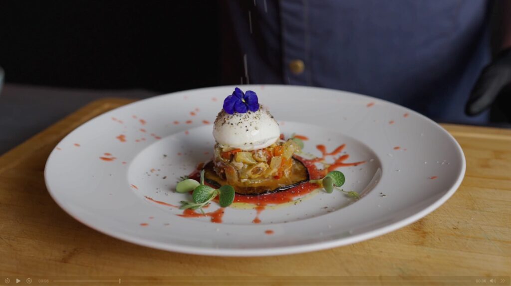 Ein plattiertes Gourmet-Gericht mit geschichtetem Gemüse, einem pochierten Ei obenauf, garniert mit einer lila essbaren Blume und Mikrogrün, beträufelt mit Sauce auf einem weißen Teller.