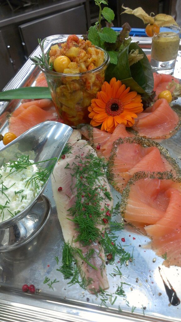 Eine Platte mit geräucherten Fischscheiben, einem Glas Meeresfrüchtesalat, frischem Grün, einem Löffel Sahnesauce und garniert mit Dill und einer Orangenblüte.