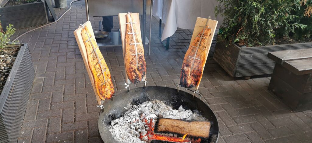 Drei Holzbretter mit Fischfilets sind vertikal um eine Feuerstelle herum angeordnet und werden über glühenden Kohlen im Freien gegart.