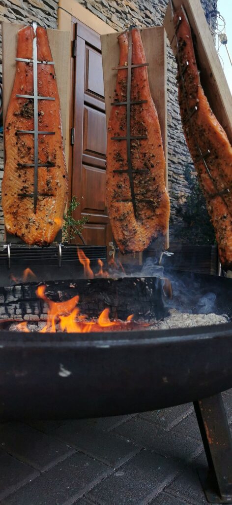 Drei große, gewürzte Fischfilets werden vertikal auf Holzbrettern montiert und über einem offenen Feuer im Freien mit einer Steinmauer im Hintergrund gegart.