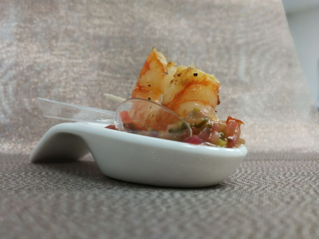 Ein Stück gegrillte Garnele, aufgespießt mit einem Zahnstocher, serviert auf einer kleinen weißen Schale mit einem Plastiklöffel und einem Topping aus gewürfeltem Gemüse, zeigt die exquisiten Leistungen der zeitgenössischen kulinarischen Präsentation.
