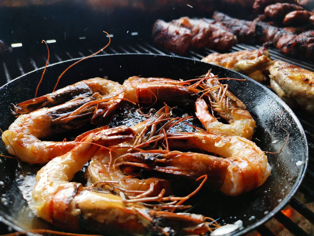 Garnelen, die in einer schwarzen Pfanne auf einem Grill gegart werden, während im Hintergrund Fleischstücke und Hähnchen gegrillt werden - ein Beispiel für die vielfältigen Leistungen des geschickten Grillens.