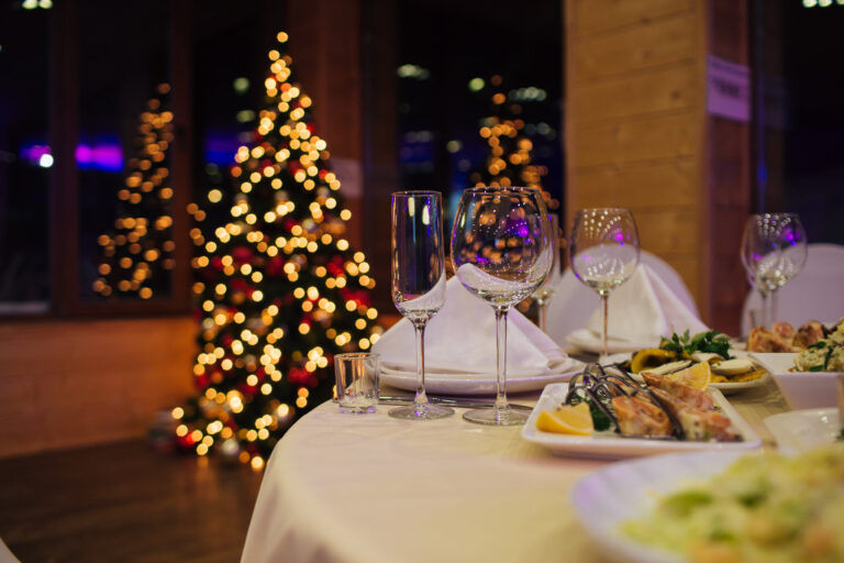 Ein geschmückter Weihnachtsbaum mit Lichtern steht an einem Fenster hinter einem runden Tisch mit Weingläsern, weißen Servietten und Tellern mit festlichen Speisen bei einem Weihnachtseventcatering im Haus.