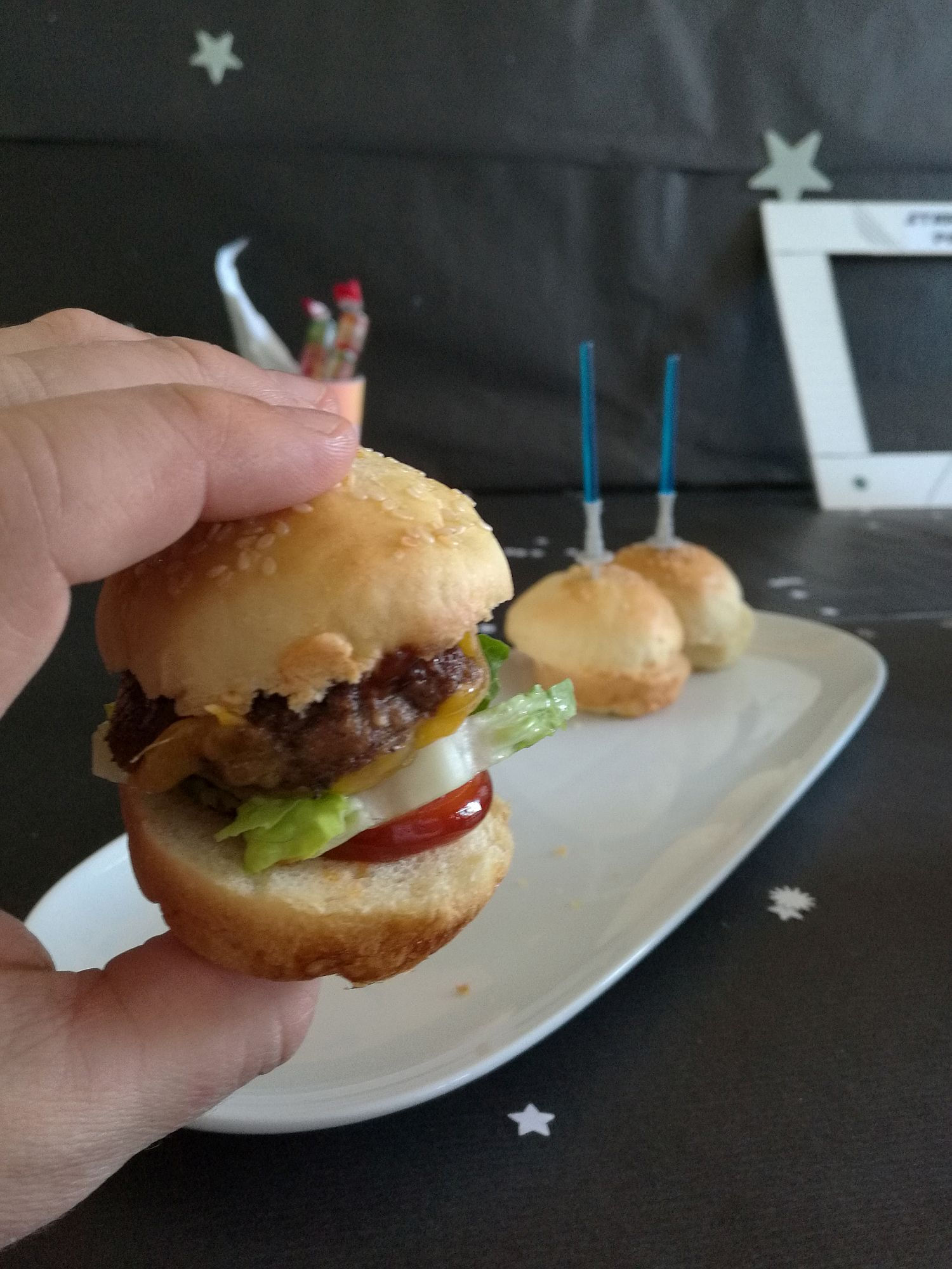 Eine Hand hält einen kleinen Hamburger mit Salat, Tomate, Käse und Rindfleischpastete; zwei weitere Slider auf einem weißen Teller liegen auf einem Tisch mit einem Sternenhintergrund.