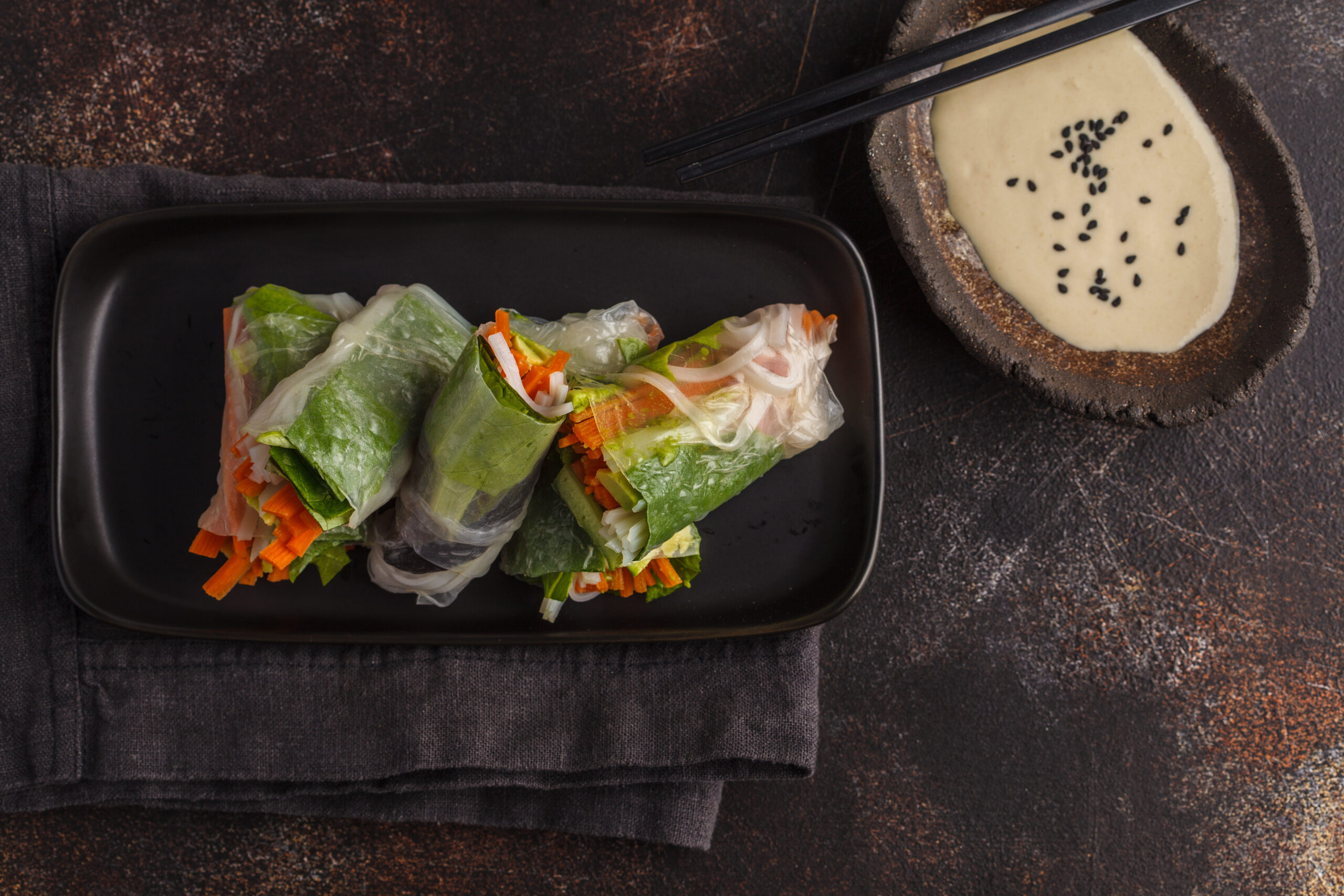 fresh handmade vegan asian spring rolls with rice noodles, avocado, carrots and tahini dressing on black dish, dark background. top view, copy space.