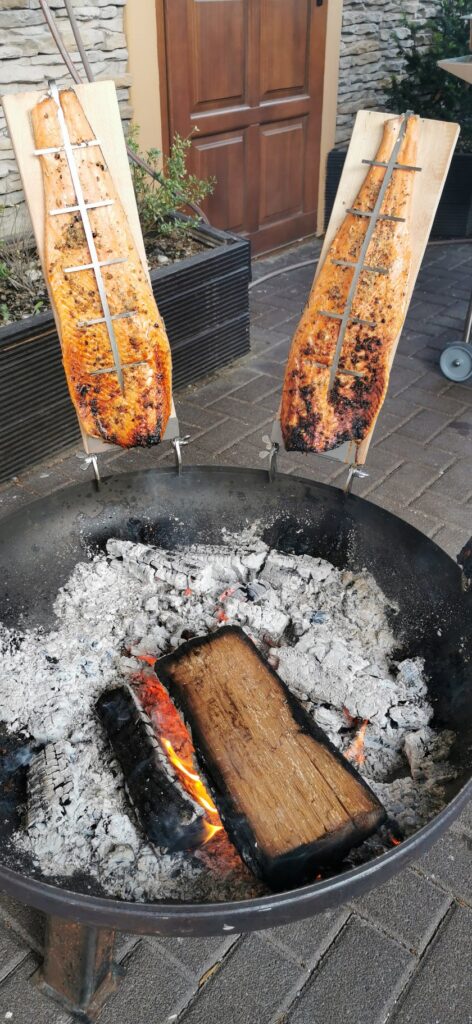 Zwei Fischfilets werden auf Holzbrettern senkrecht über einer Feuergrube mit brennendem Holz und Asche gegart, wodurch eine rustikale Szene entsteht, die an die traditionellen Mollerhaus-Treffen im Logensaal erinnert.