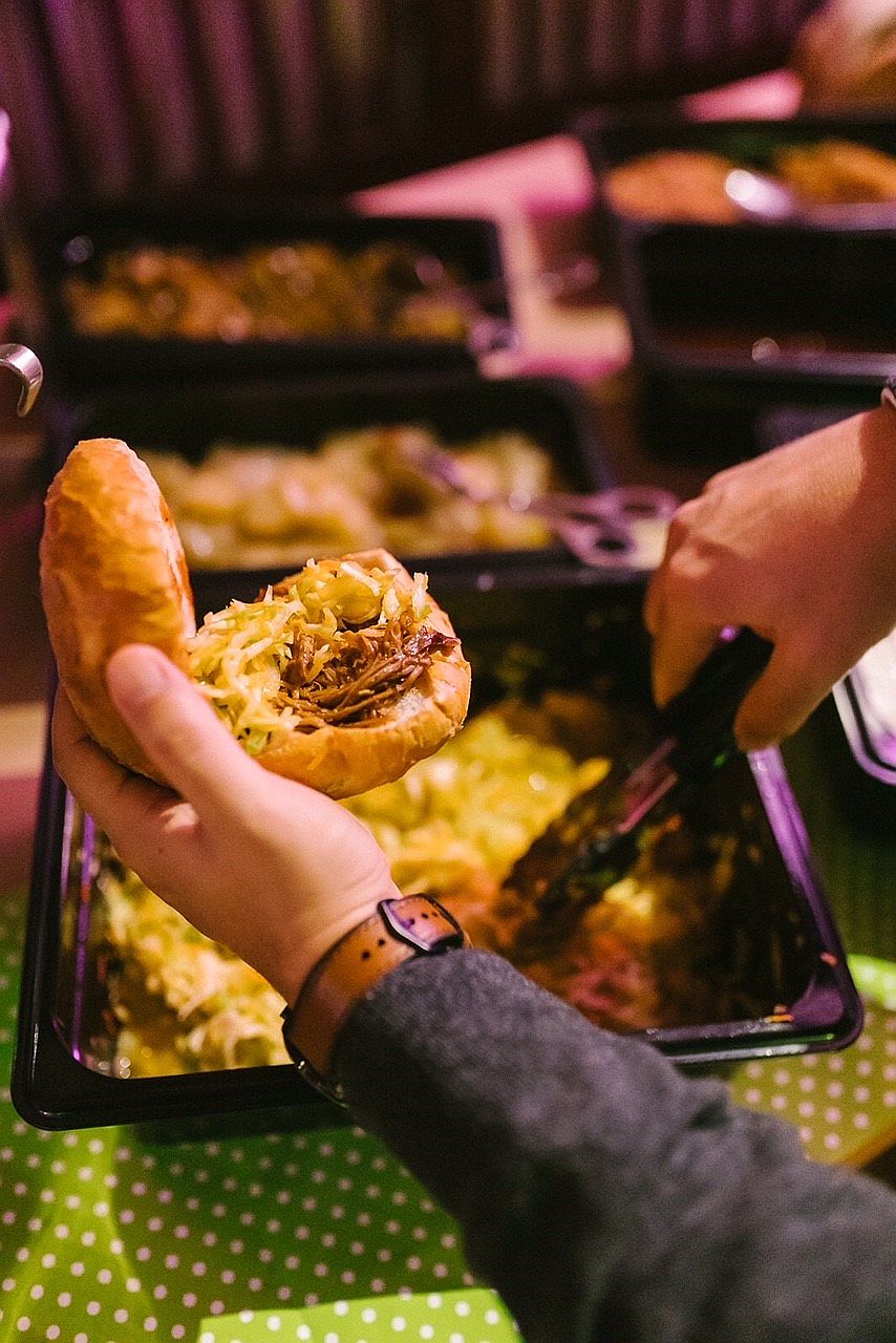 Eine Person benutzt eine Zange, um ein Sandwich über einem Tablett mit Speisen an einem Buffet mit mehreren Tabletts im Hintergrund zu belegen.