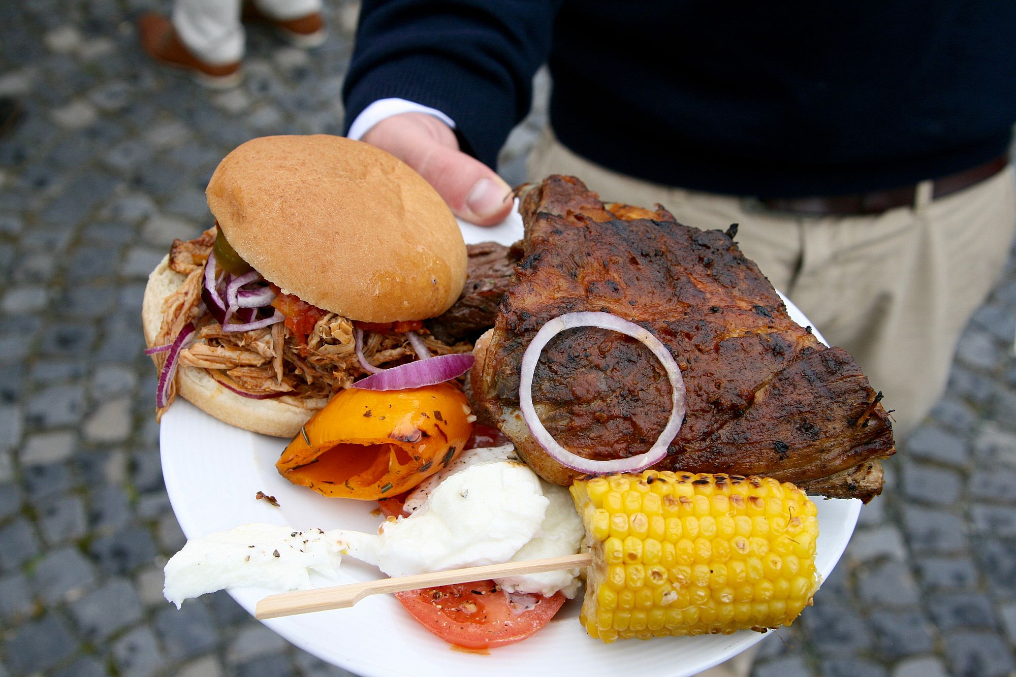 Eine Person hält einen Teller mit gegrilltem Steak, einem Pulled-Pork-Sandwich, Maiskolben, gegrillter Paprika, Zwiebeln, Tomatenscheiben und einer Kugel Weißkäse.