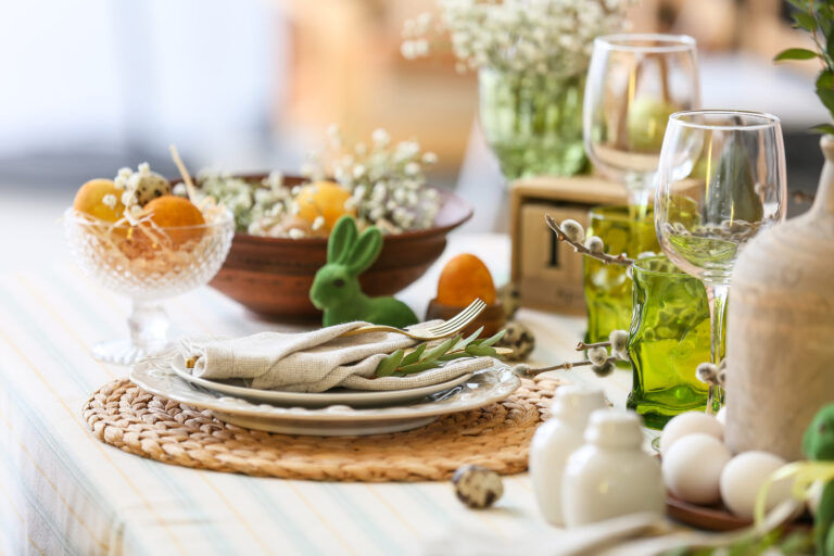 Eine vom Frühling inspirierte Tischdekoration mit gestapelten Tellern, verschachteltem Besteck, grünen Gläsern, Eiern, dekorativem Grünzeug, einer Hasenfigur und einer Obstschale auf einem gewebten Tischset - perfekt für das Ostercatering.